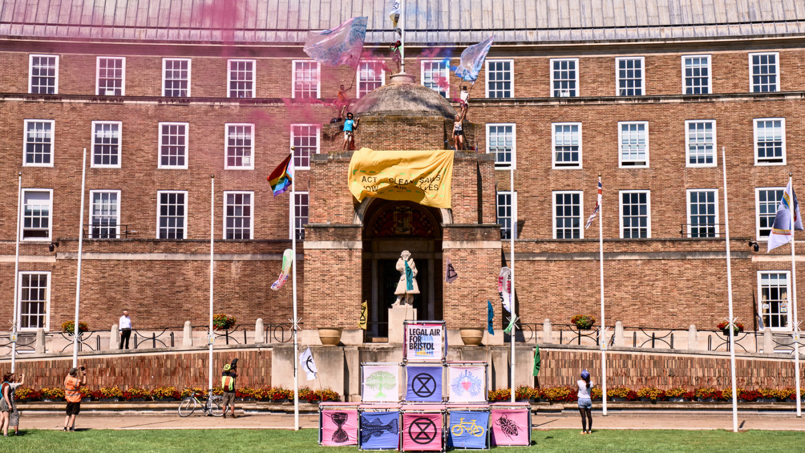 Photo: Extinction Rebellion wave flags atop the roof of City Hall amidst colourful smoke from smoke bombs and a sign that imitates coronavirus messaging, saying 
