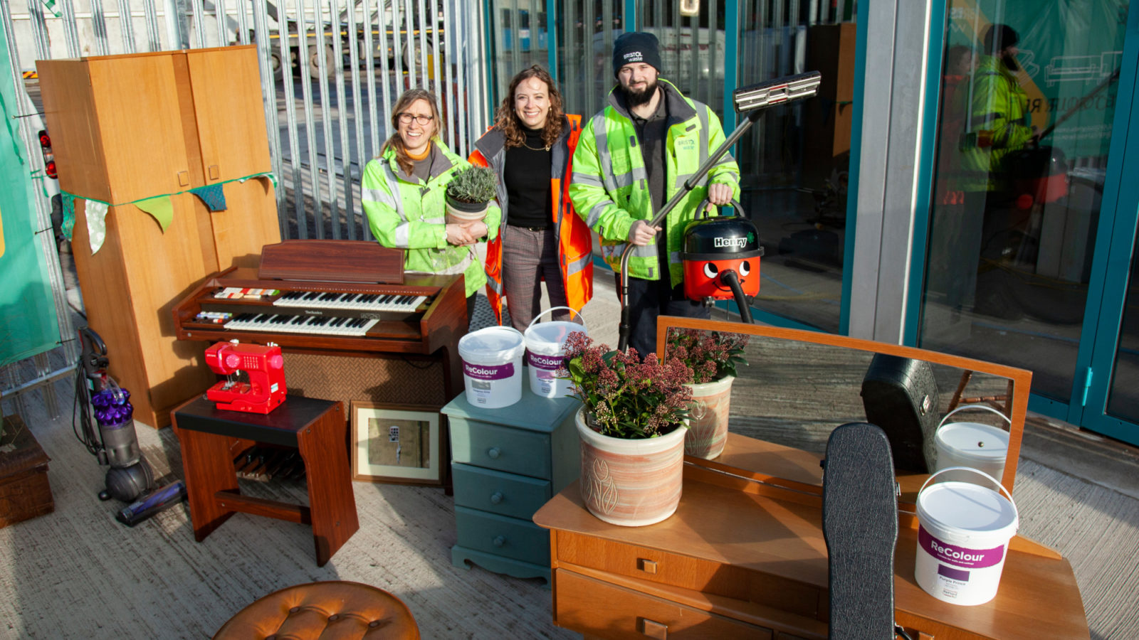 Photo: Assistants stand behind a number of items including a cupboard, a music keyboard, a sewing machine, and a stool at Bristol Waste's reuse waste shop