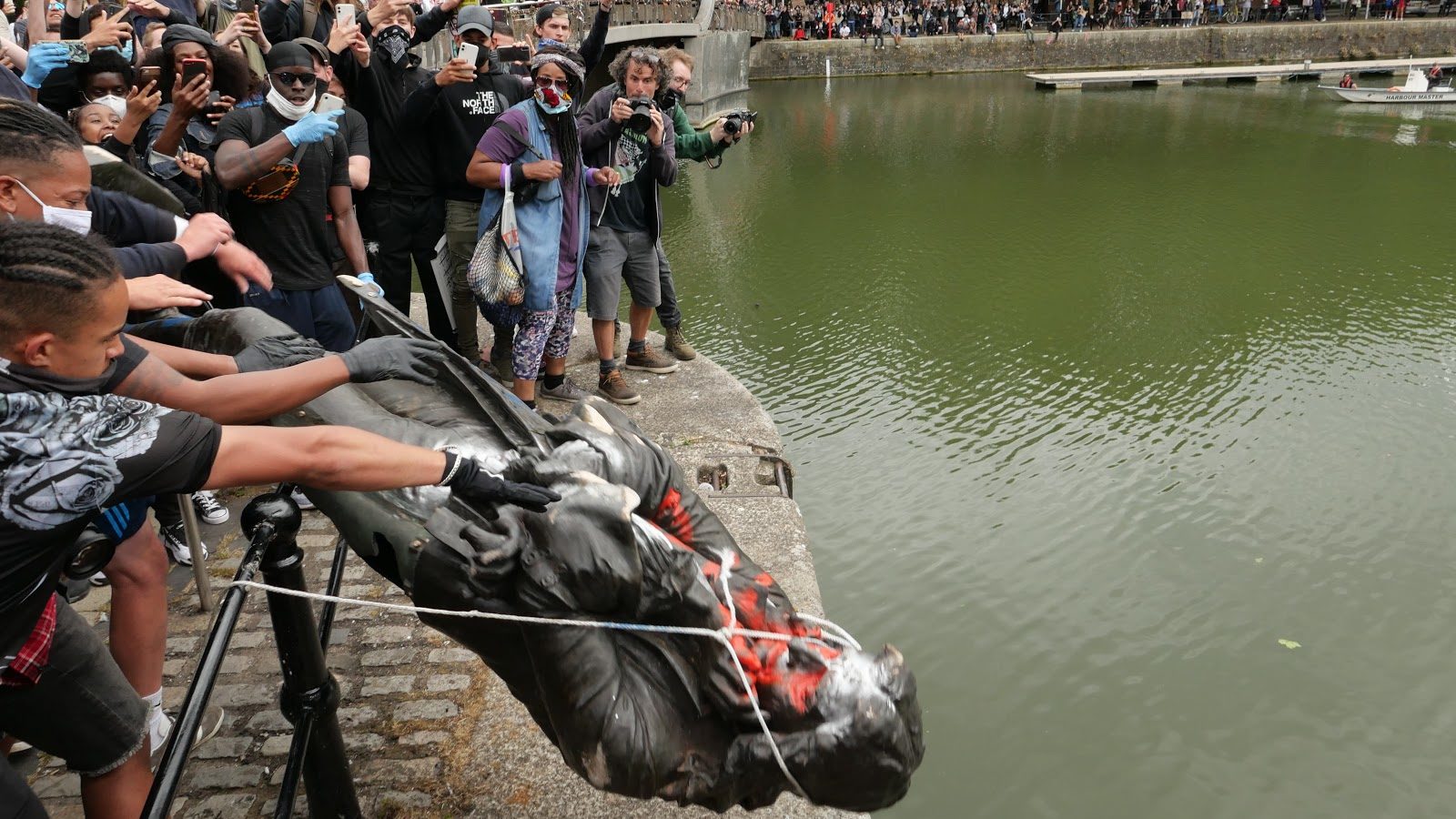 Photo: Crowds gather around the Floating Harbour and stand on Pero's Bridge as the statue of Edward Colston, covered in graffiti and with a rope around its neck is lifted over a fence to be thrown in to the water below.