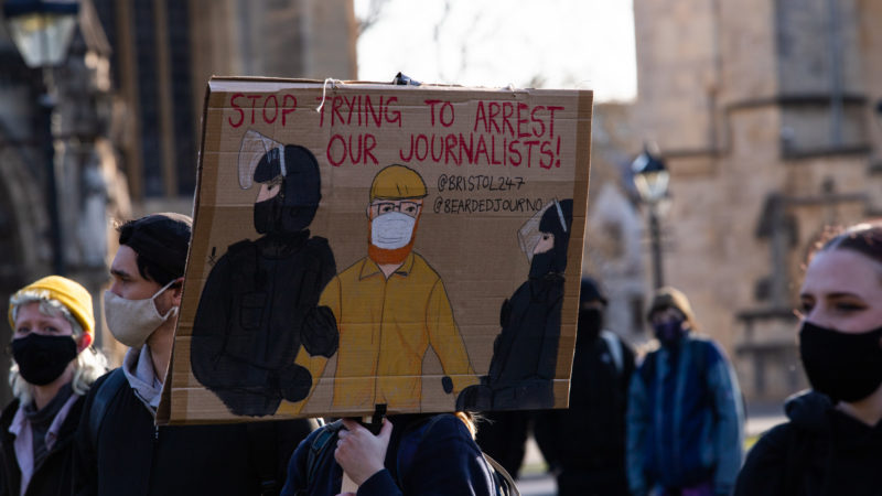 A person holds a cardboard banner reading 'stop trying to arrest our journalists'