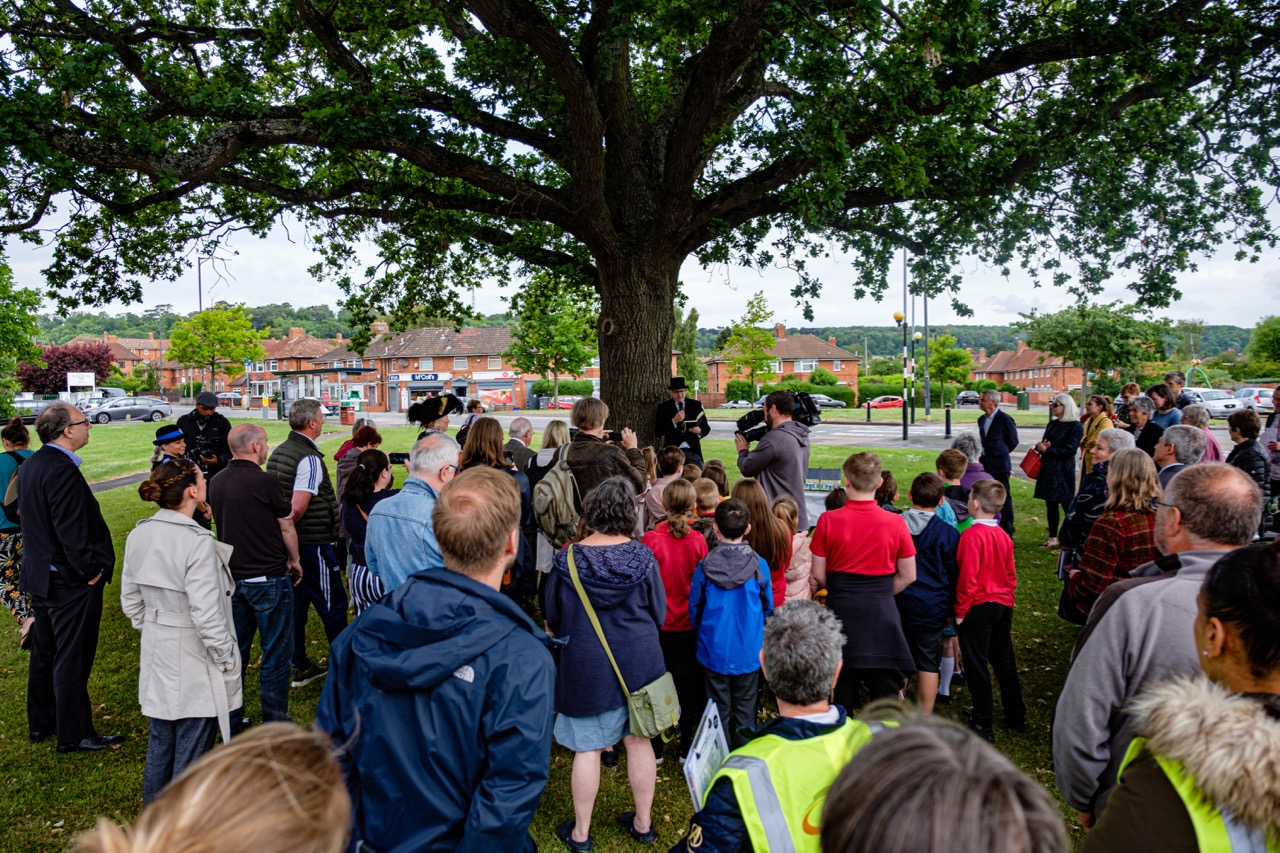 Group of people including school children gathered under a large tree, a TV crew is also in attendance