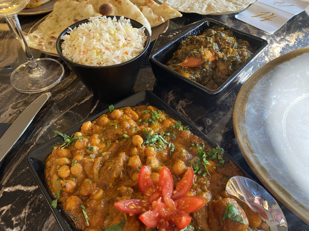 A plate of Afghan lamb curry in the foreground, with rice and saag aloo behind.