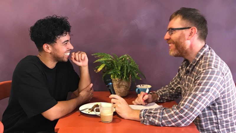 Martin Booth interviews a young man over a cup of coffee