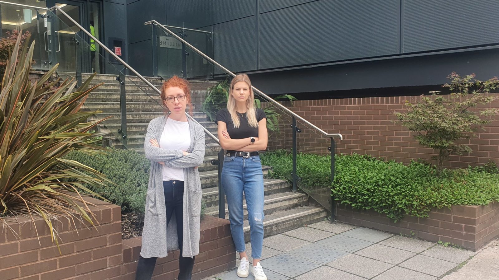 Blonde woman in 20s stood looking unhappy with arms folded in front of tall building