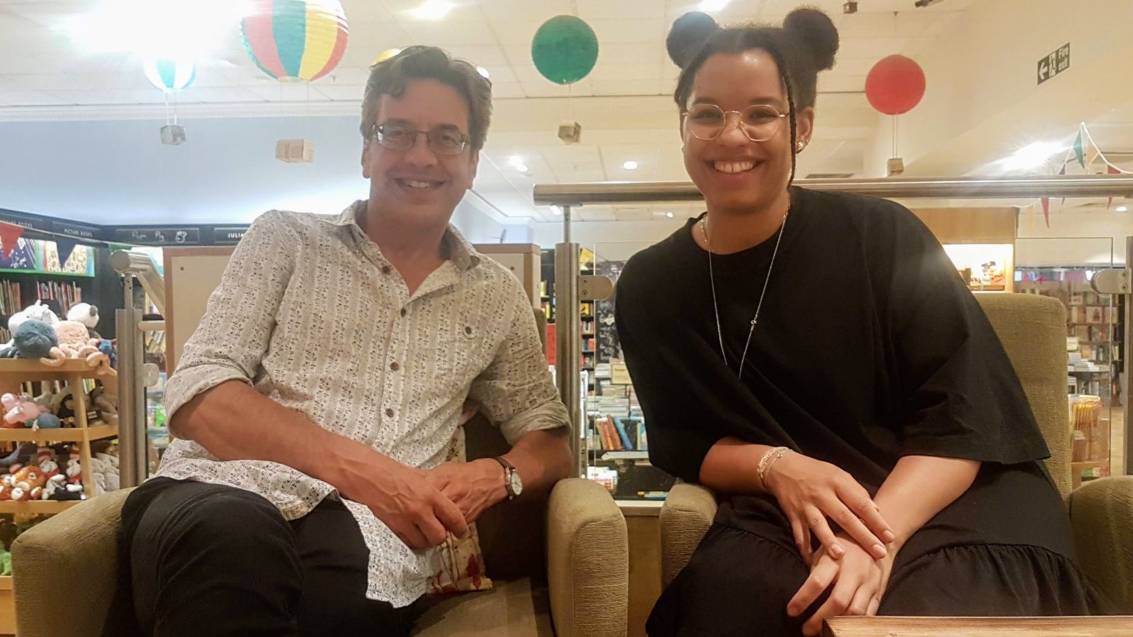 A man and a woman sit side by side in a bookshop facing the camera.