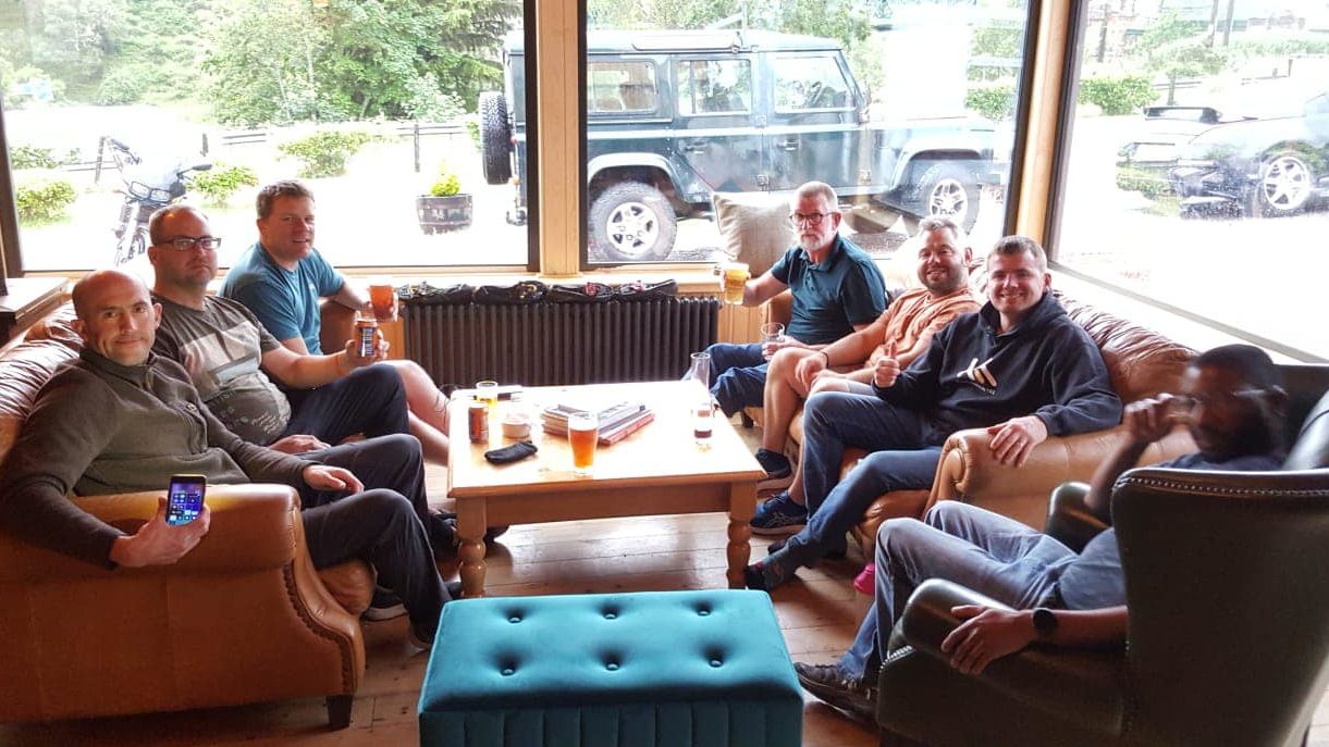 6 men sit on brown leather sofas in a bright window lit room, they are relaxing after a long cycle.