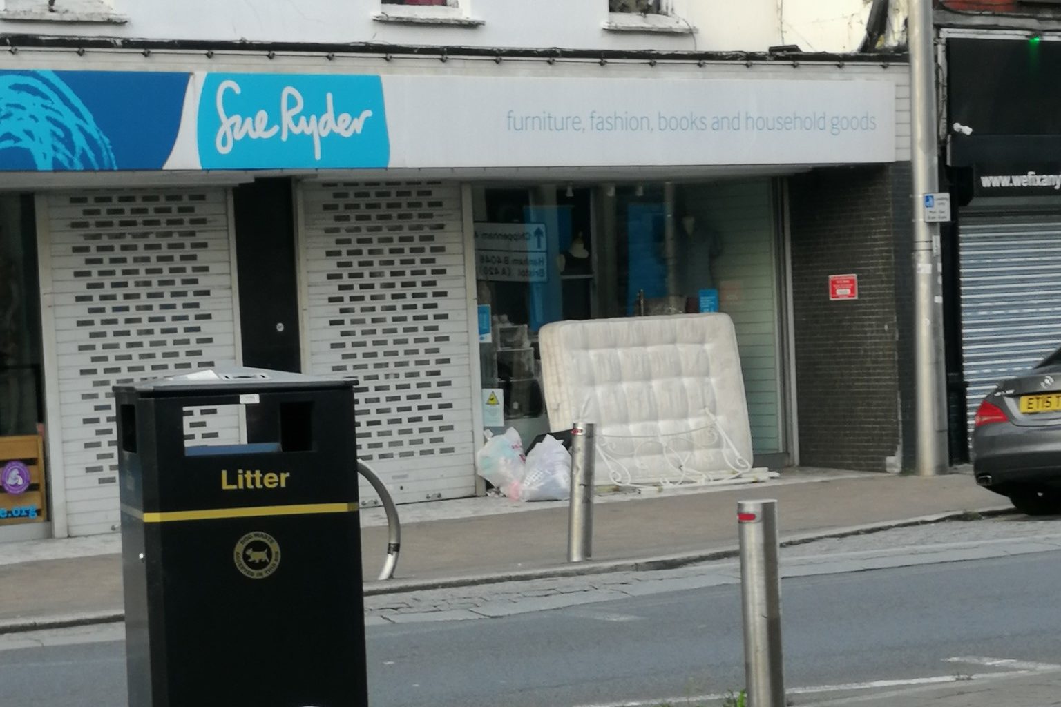 A mattress and metal headboard are leant against the window of Sue Ryder charity shop. Next to it are several bags of clothes which have been ripped open, clothes are all over the entrance to the shop and the pavement. The shop is clearly closed as the security grating is down.