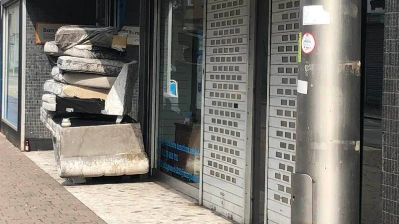 An upside down sofa with sofa cushions piled on top of it sits under the awning of a shop, the shop has its security grates down and is clearly closed. The sofa itself is dirty and wet.