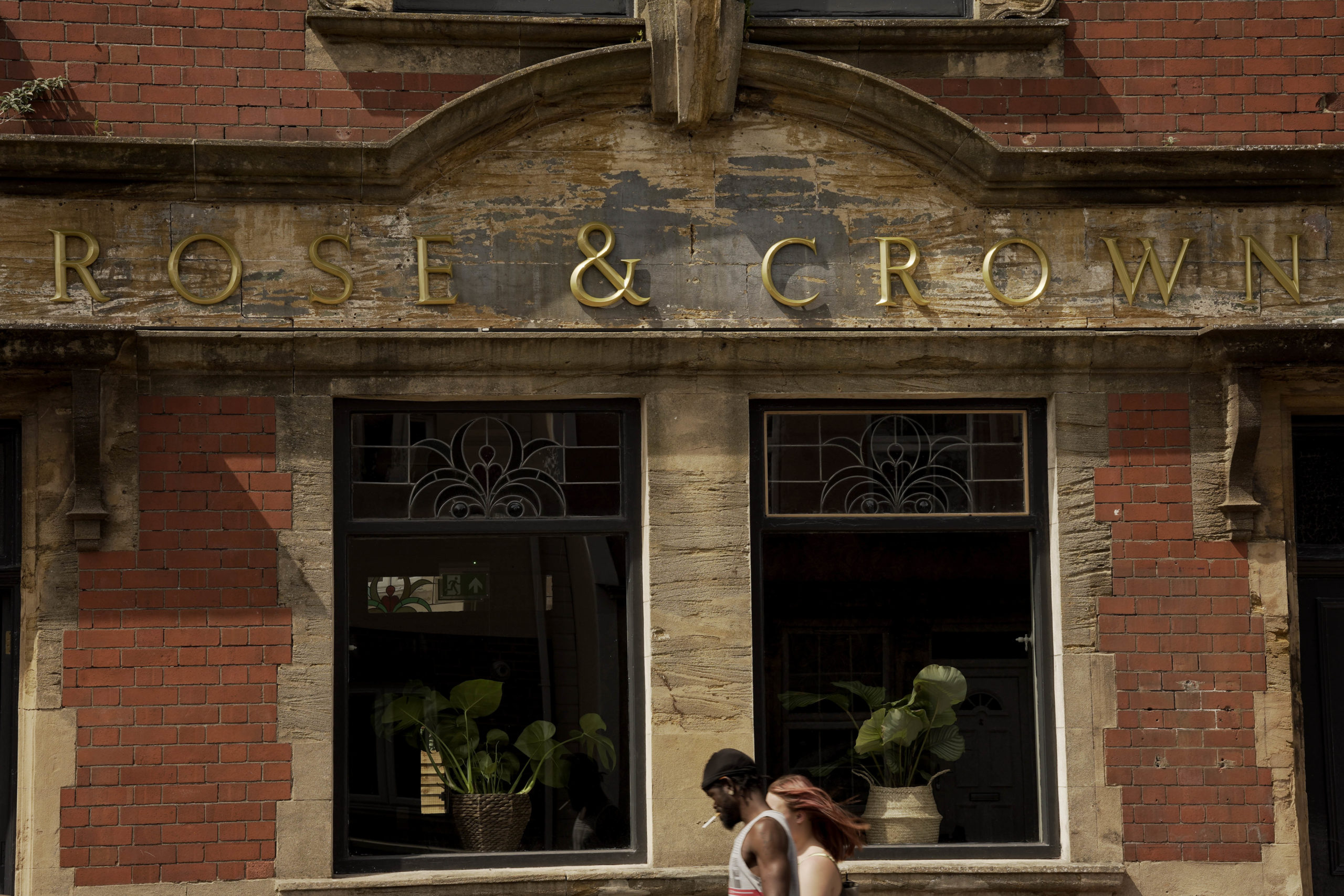 The front of the Rose & Crown, a red brick pub. "Rose & Crown" is written in gold letters, stuck on across the front entrance. In the windows there are lots of green plants in pots. 