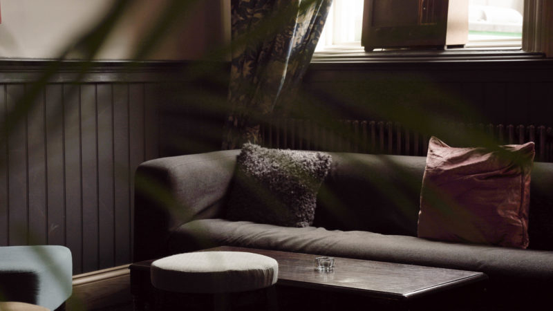 A grey sofa underneath a big window, the sofa has several pillows and a dark wood table in front of it. 