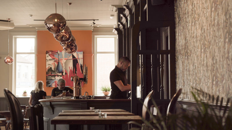 A wide angle of the pub, showing the bar and the barman getting change from the till. There are big orange globe lights above the bar and in the background you can see people sitting at tables chatting and enjoying a drink.