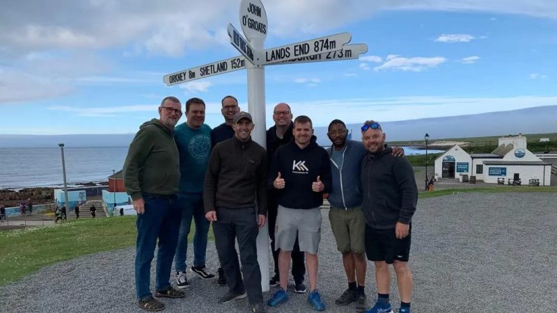 6 men at the John O'Groats which points to Land's End. It's foggy, they are smiling. photo Trevor Wilson
