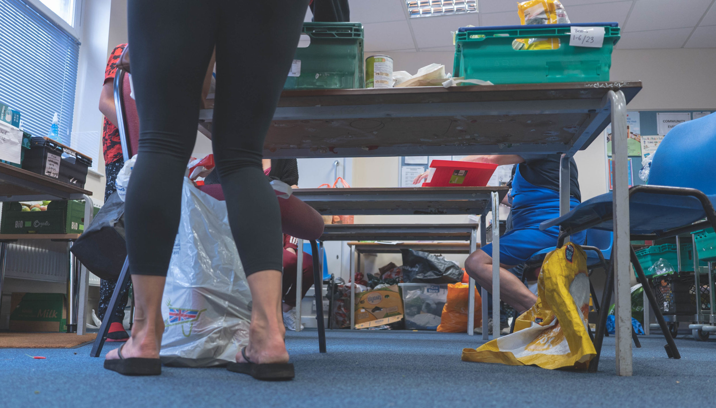 Taken from a low angle, you can see the legs of two food bank users and a volunteer. One client and volunteer are sitting at opposite ends of a table talking. The other client is standing next to another table which has a box of food, they are putting the food into bags. You can’t see any of their faces, the shot is cut off at their legs.