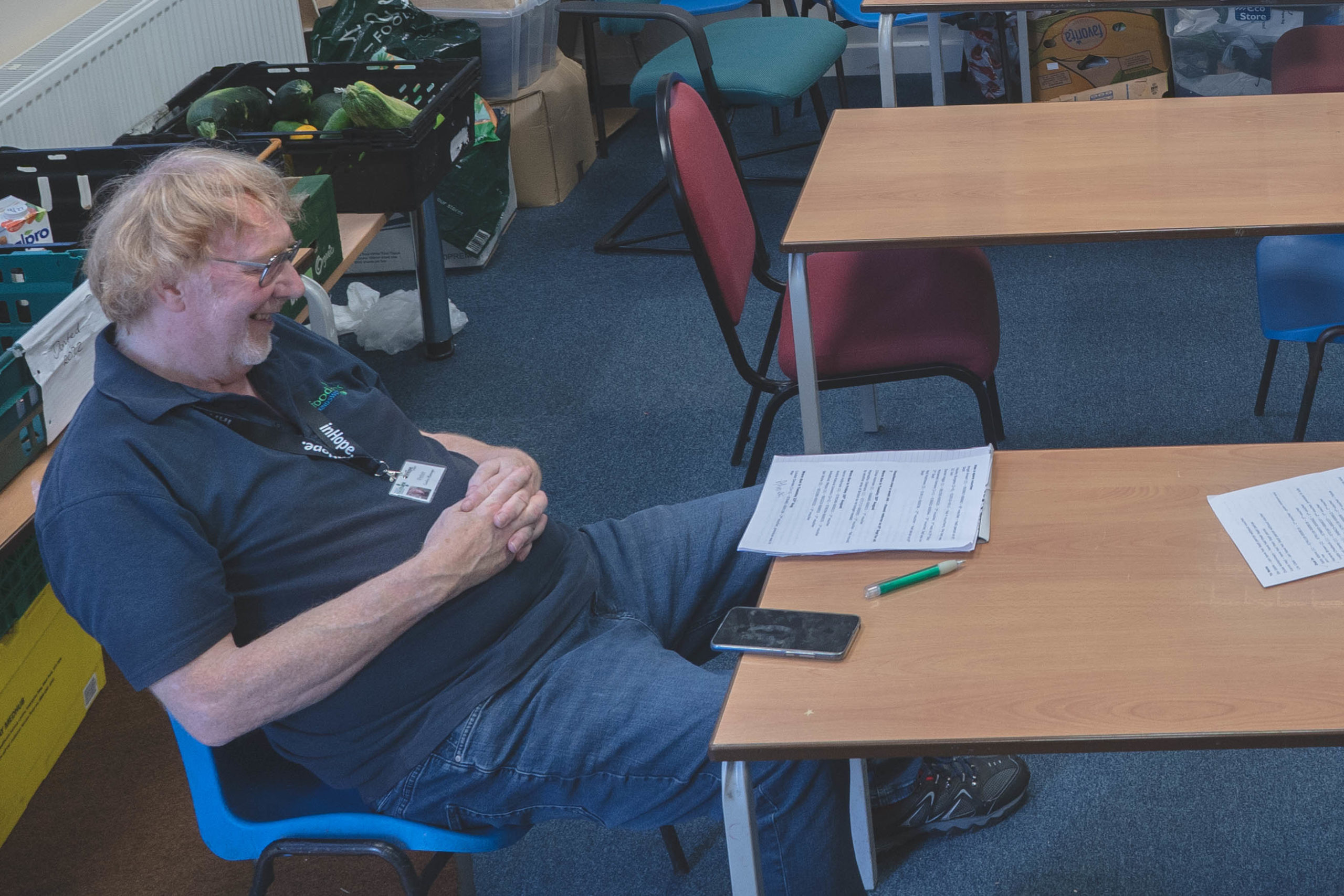 Peter Shears, wearing his blue Trussell Trust polo shirt, sits in a blue plastic chair and rests with his hands on his stomach. He is smiling but looks tired. There are papers and his phone on the desk in front of him.