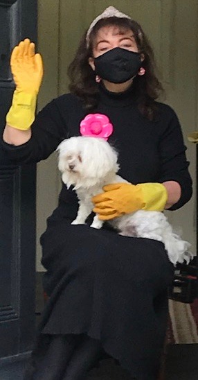 Jennifer, a white woman with dark curly hair is sitting in her front door, with a small white dog on her lap. She's wearing pink kitchen gloves and a mask. 