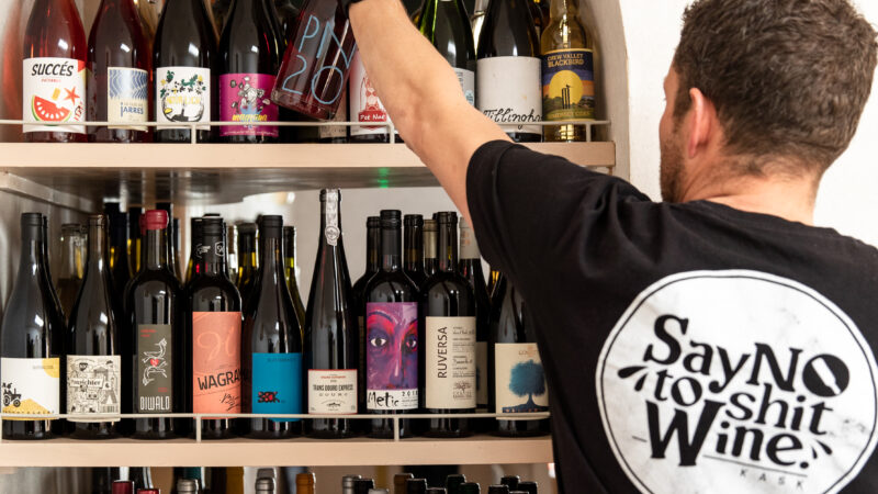 A man reaches for a bottle of KASK wine from a shelf. His t shirt reads 'say no to shit wine'