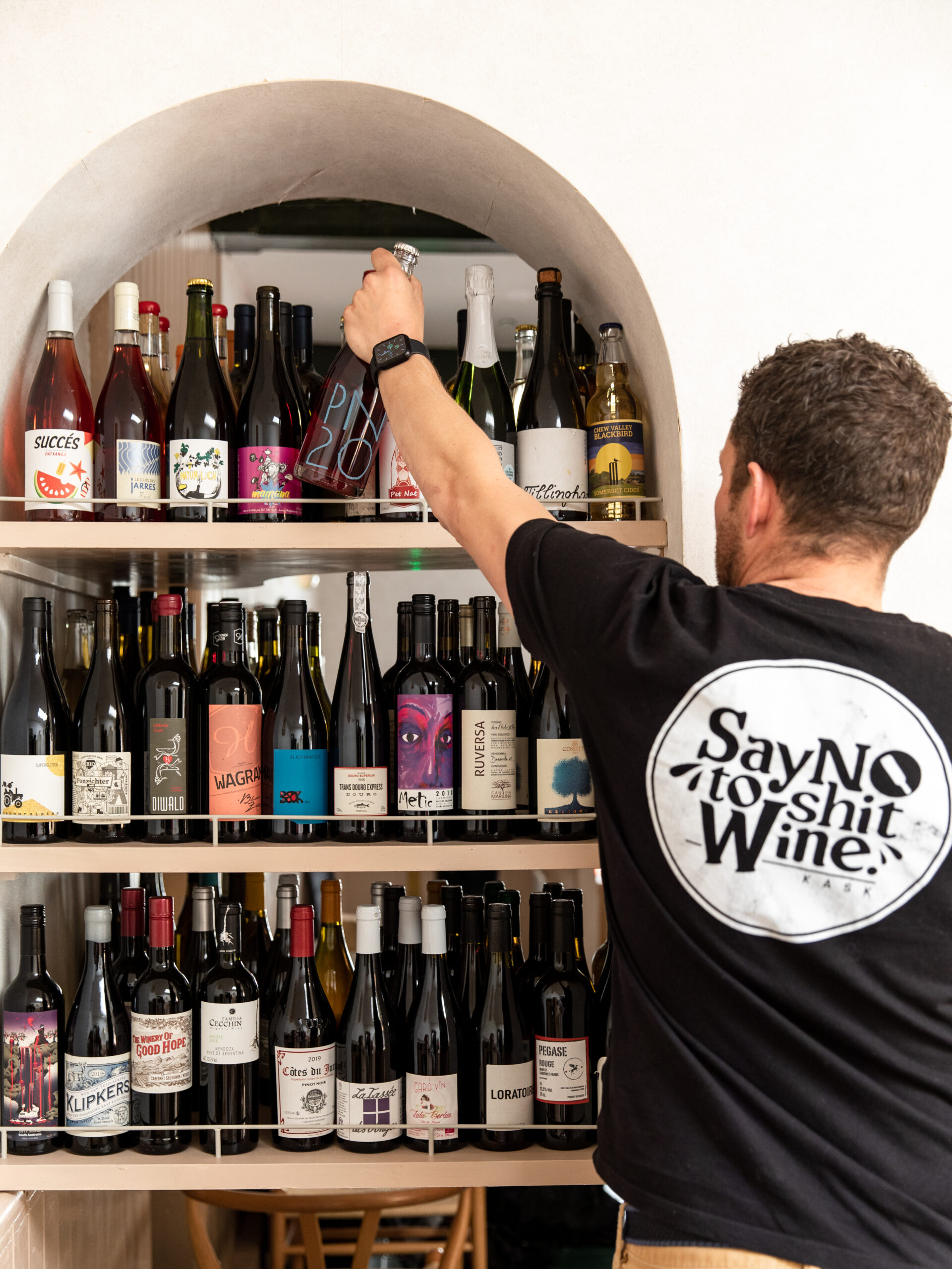 A man reaches for a bottle of KASK wine from a shelf. His t shirt reads 'say no to shit wine'