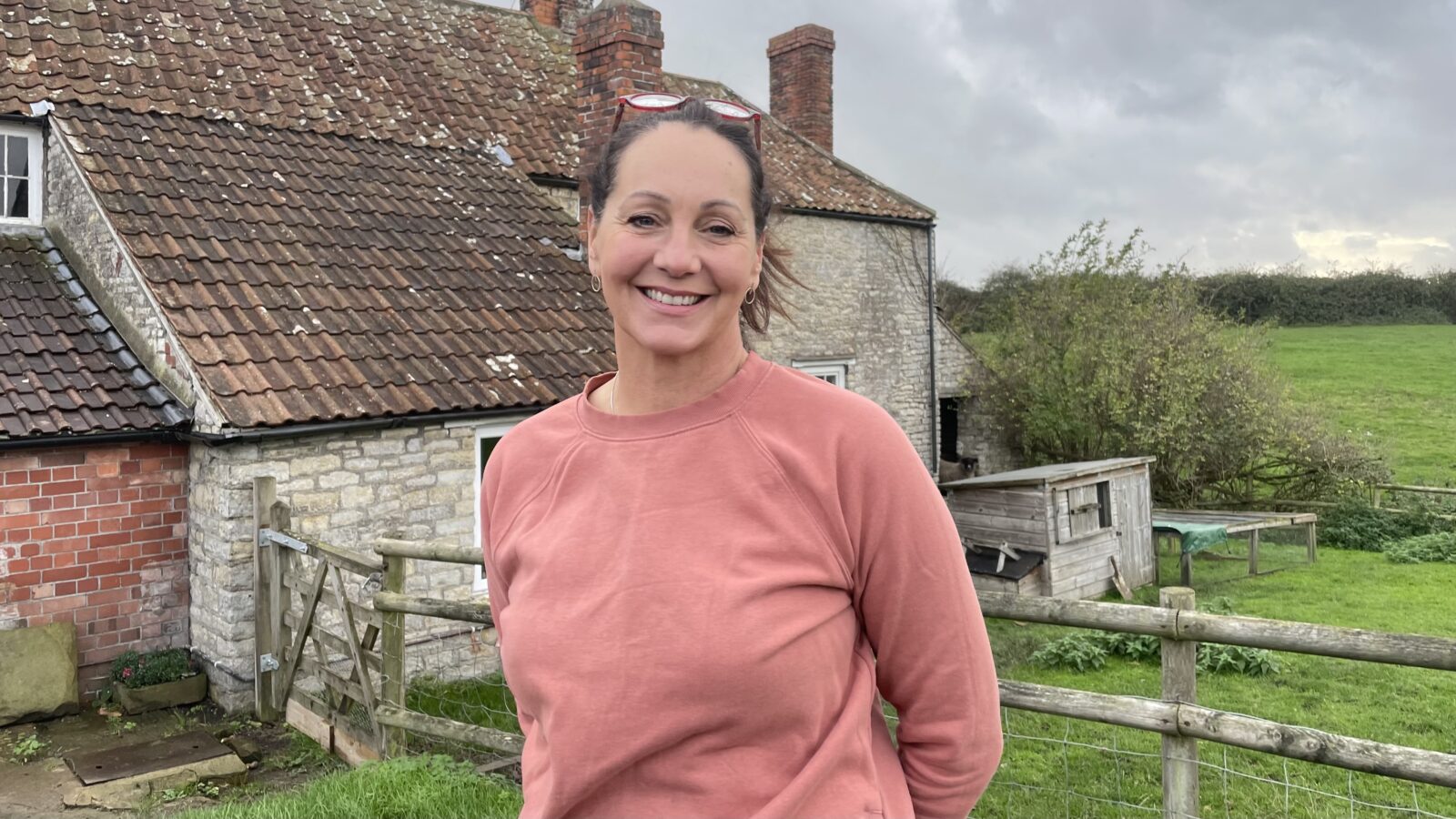 Farmer Catherine Withers waring a light pink top smiling outside the farmhouse at Yew Tree Farm, with a sheep behind her left shoulder and fields in the distance