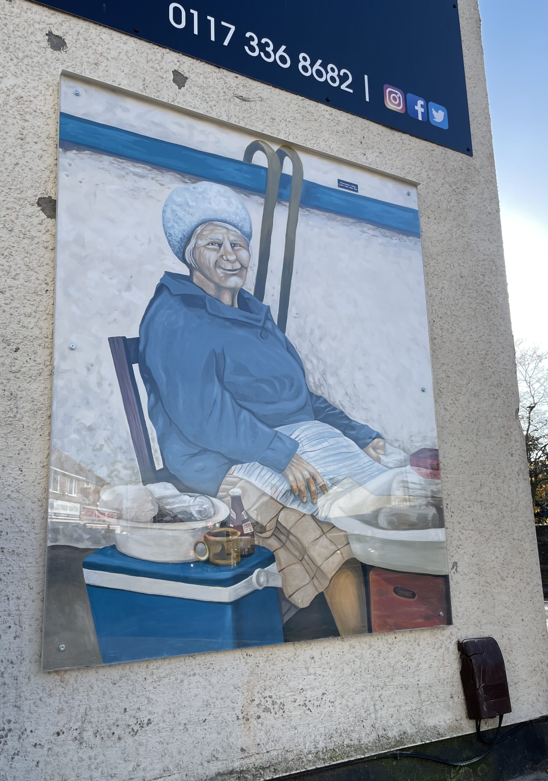 An old lady wearing a woollen hat and blue jacket sits on a chair, with two walking sticks hanging behind her