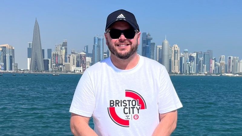 Downsy wearing a white t-shirt with Bristol City written on it, with skyscrapers on the horizon behind him