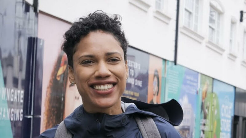 A woman stood smiling dressed in a black coat on a bicycle