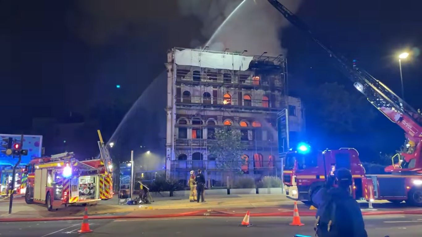 Flames are inside the Grosvenor Hotel as firefighters spray water and bystanders watch on