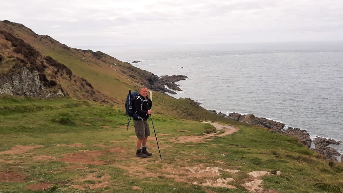 A man stood on a coastal path dressed in hiking gear on an overcast day