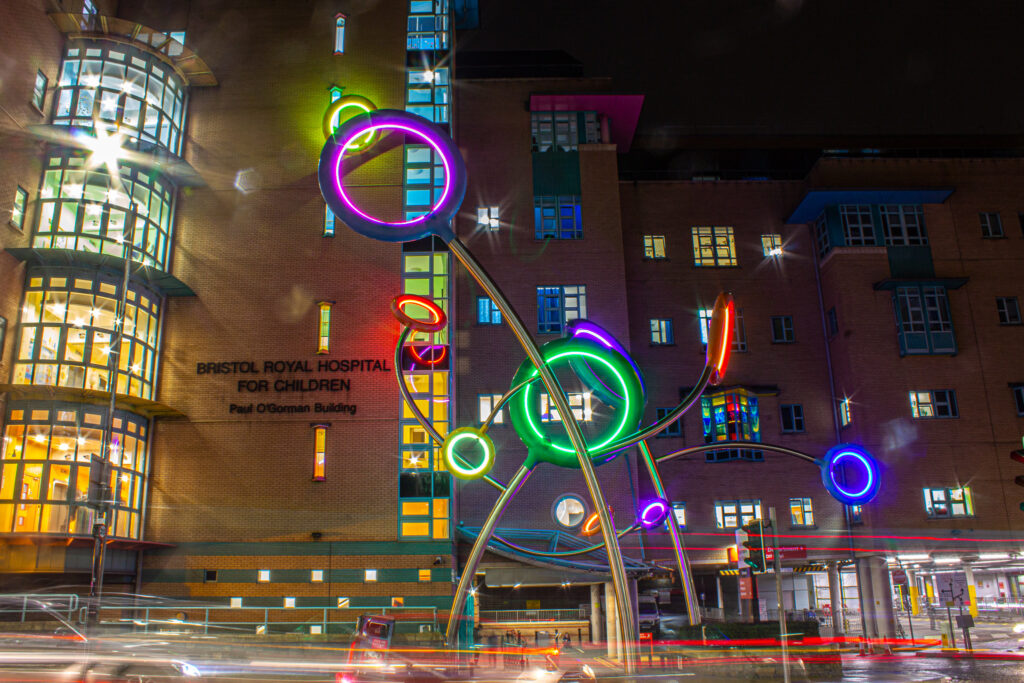 Lollipop Be-Bop sculpture outside Bristol Children's Hospital at night with cars driving by, featuring hoops lit up yellow, green and blue