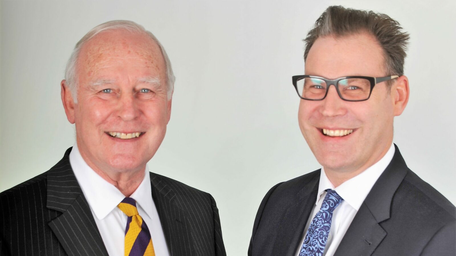 Two men, a father and a son, stood side by side smiling dressed in shirts, ties and black blazers
