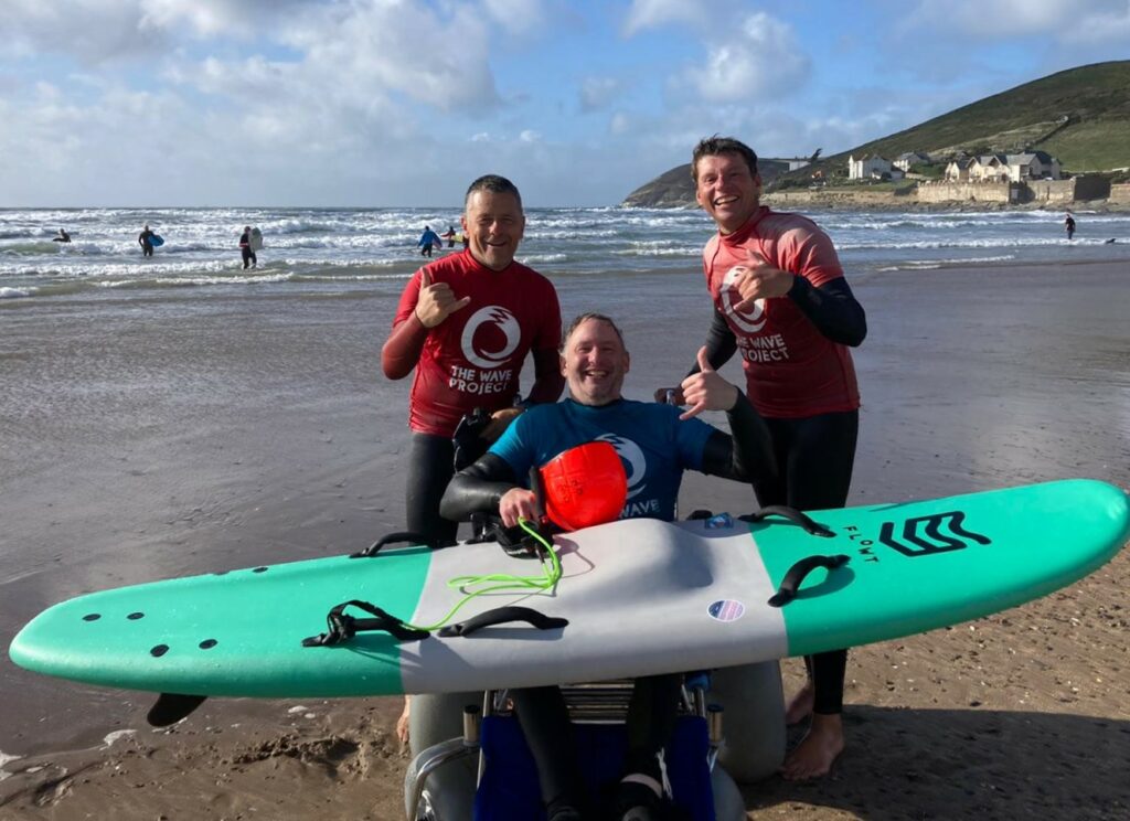 Stephen Lightbown sits in an adaptive wheelchair on the beach with a surboard balanced on his legs, with two men to either side of him both smiling