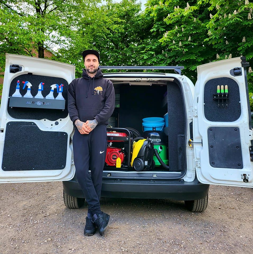 A man stood grinning in front of his van with both of the back doors open showing cleaning equipment
