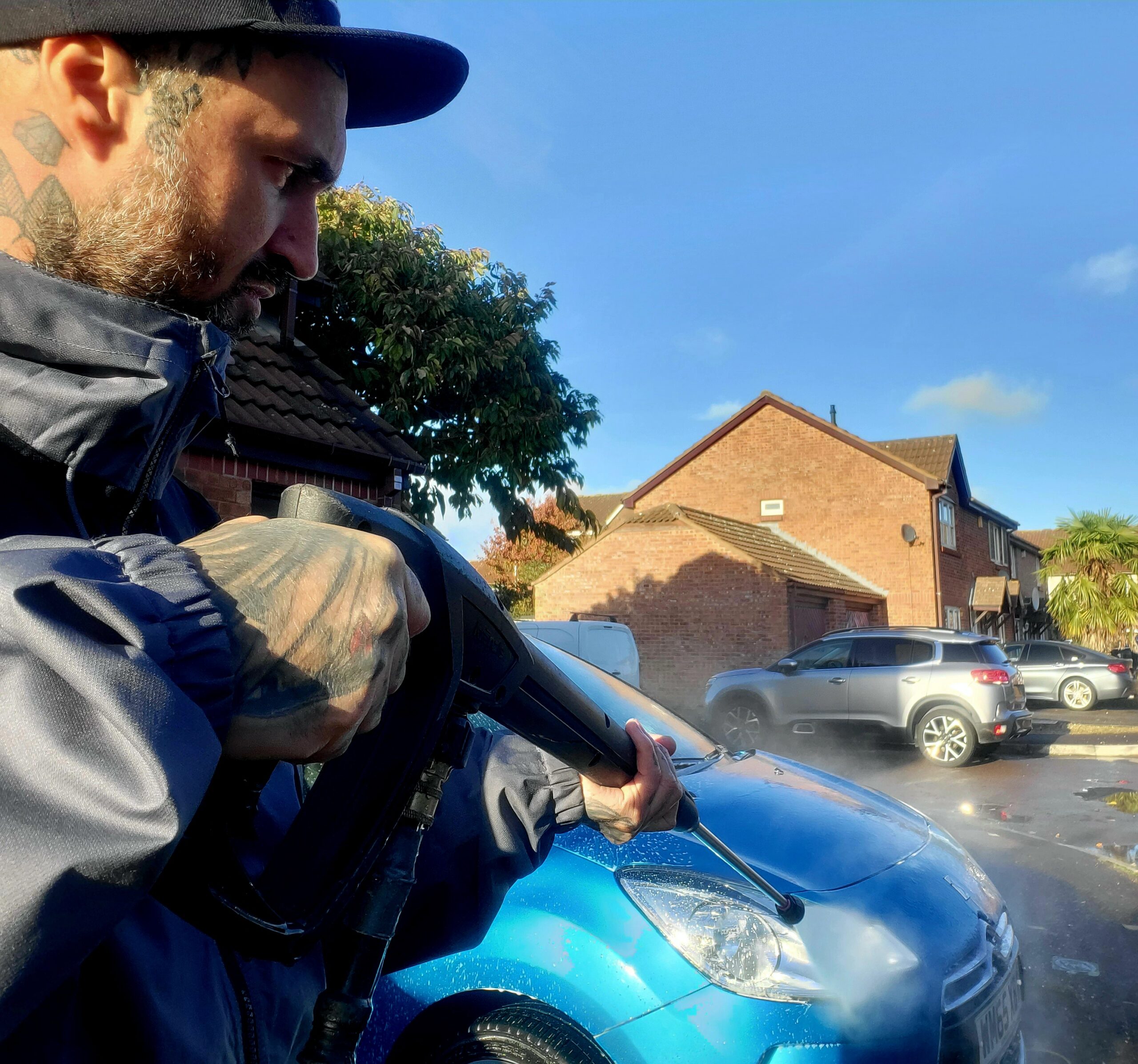 A man holding a jet wash and cleaning a car on a bright sunny day