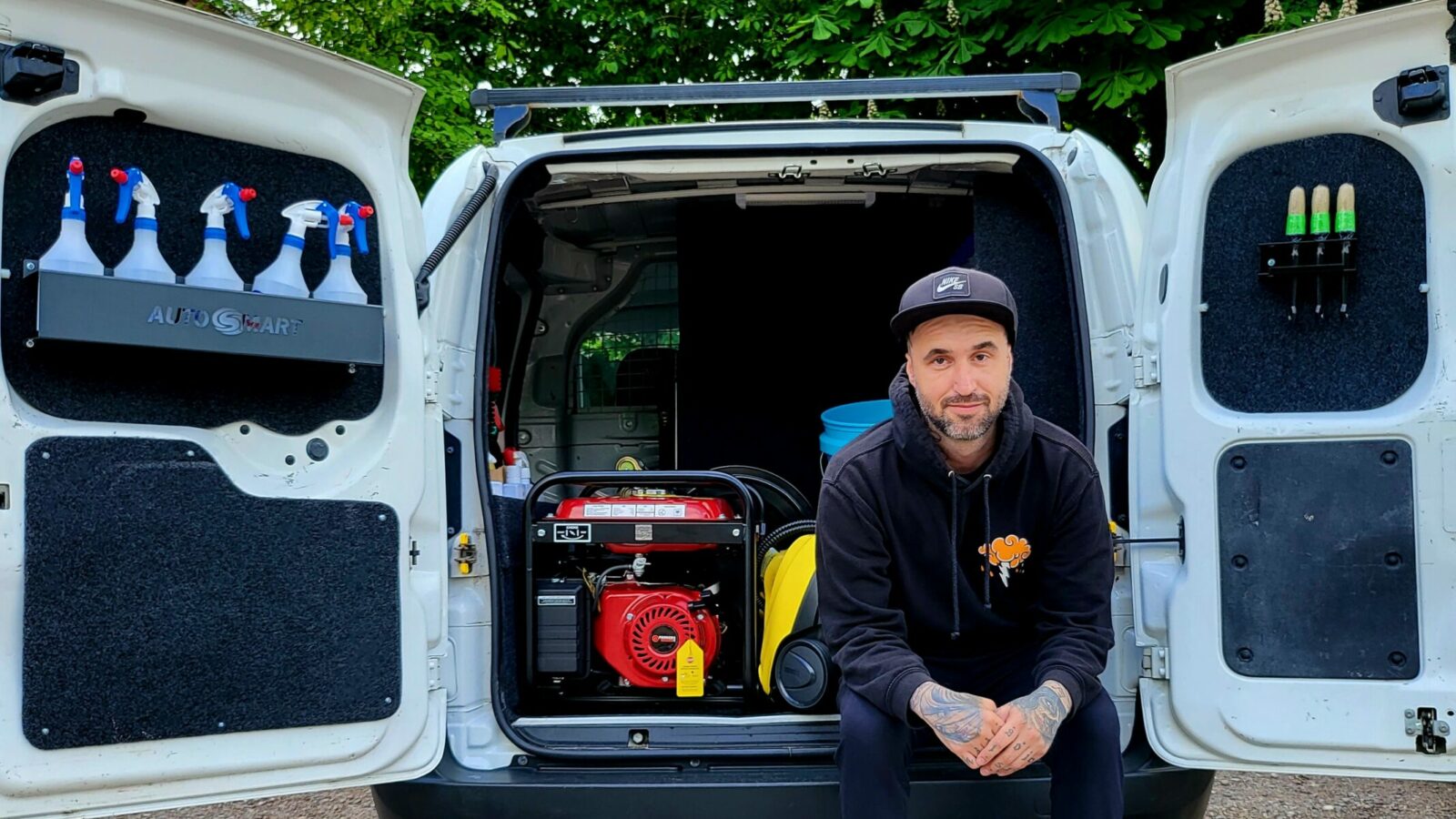 A man grinning sat inside his van with both of the back doors open showing cleaning equipment