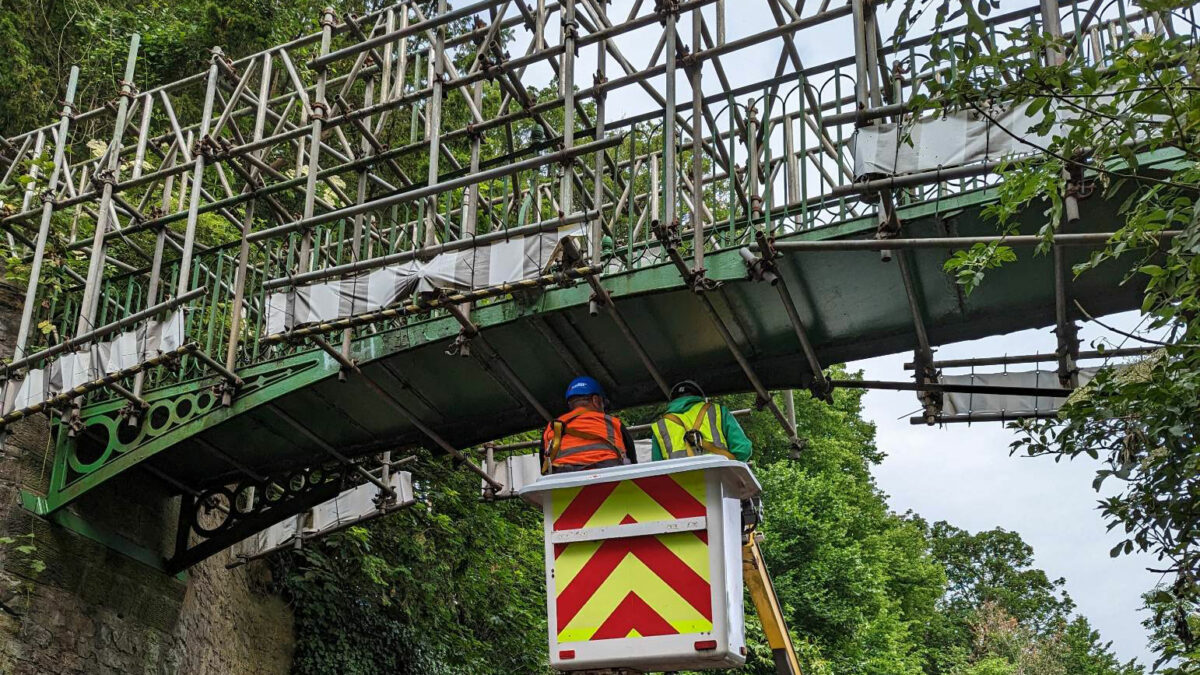 Two men in hi-vis use a platform lift to inspect the underside of a green bridge