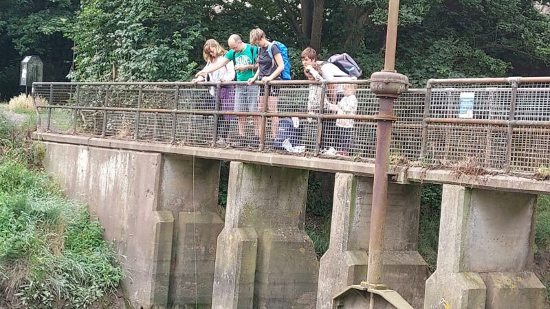 A group of people lean over a bridge. They are scooping up water from the river below, using a bottle on a long string