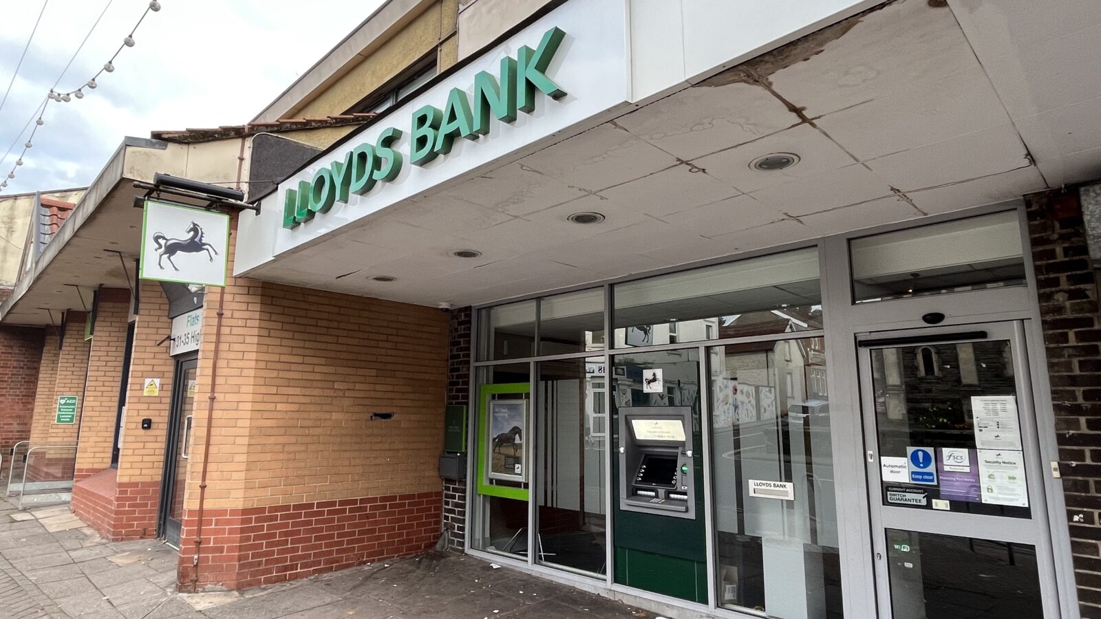 A small branch of Lloyds bank, with a green sign and a black horse logo. It has a cash point outside