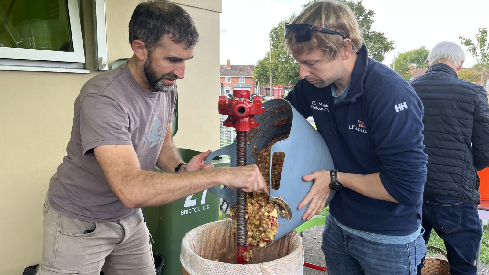 Two men pour chopped apples into a small apple press