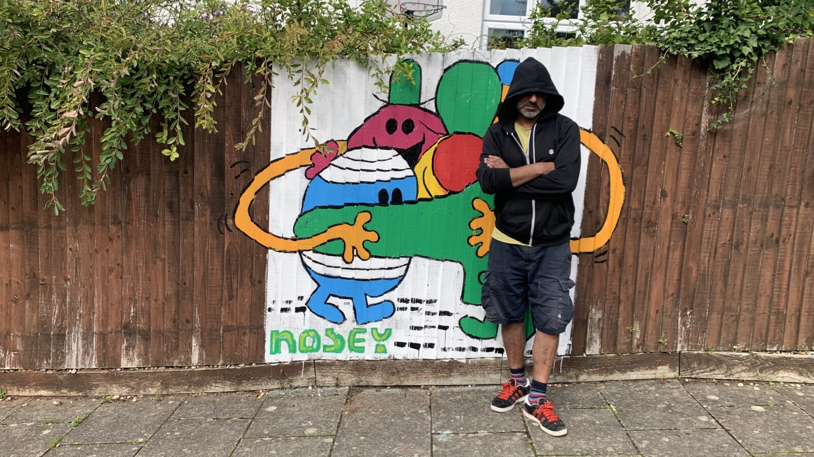 a man with a hoodie pulled over his face stands in front of a graffiti of a group of Mr Men