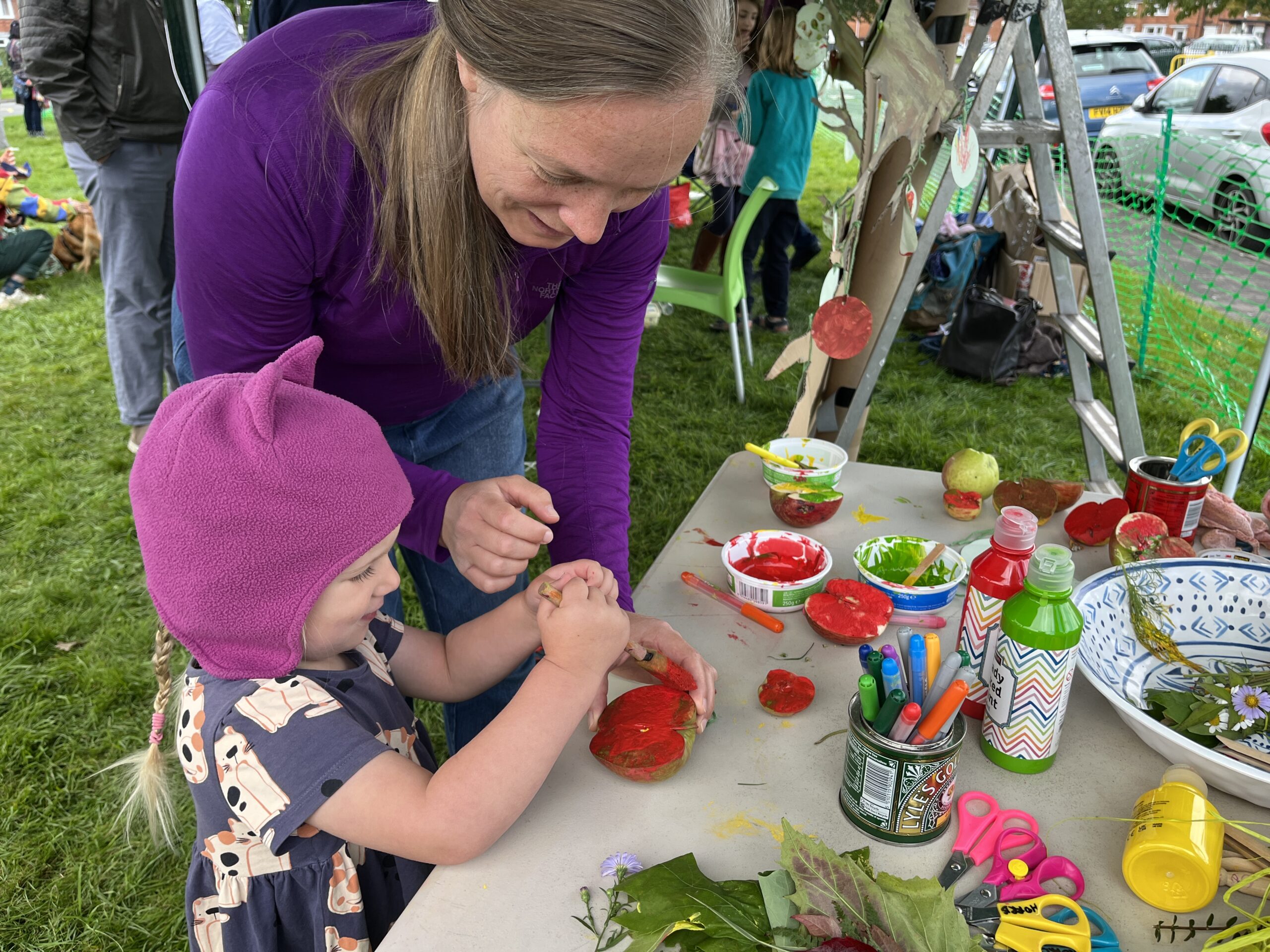 A woman helps a small child paint red paint onto half an apple
