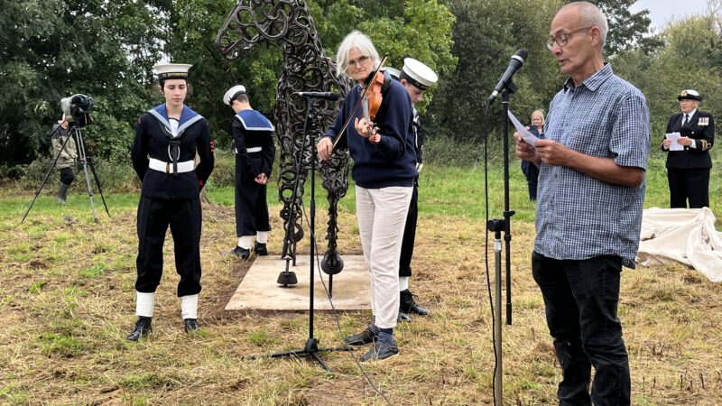 A man sings into a microphone. Accompanied by a lady on guitar