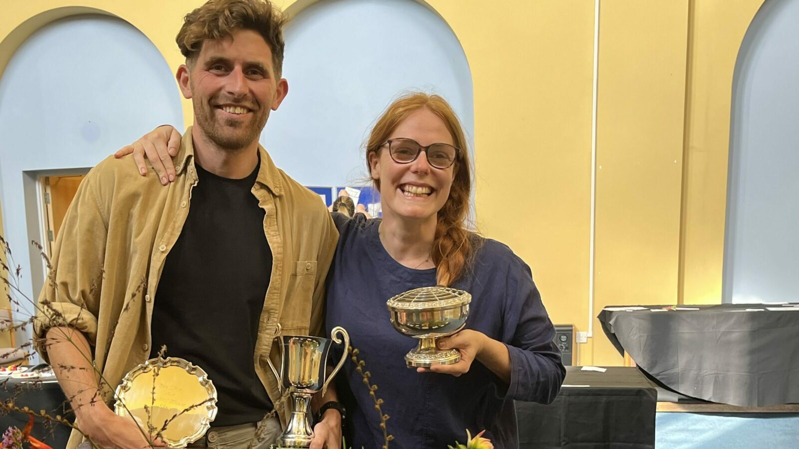 a couple stand in front of a table of flower arrangements, holding 3 trophies