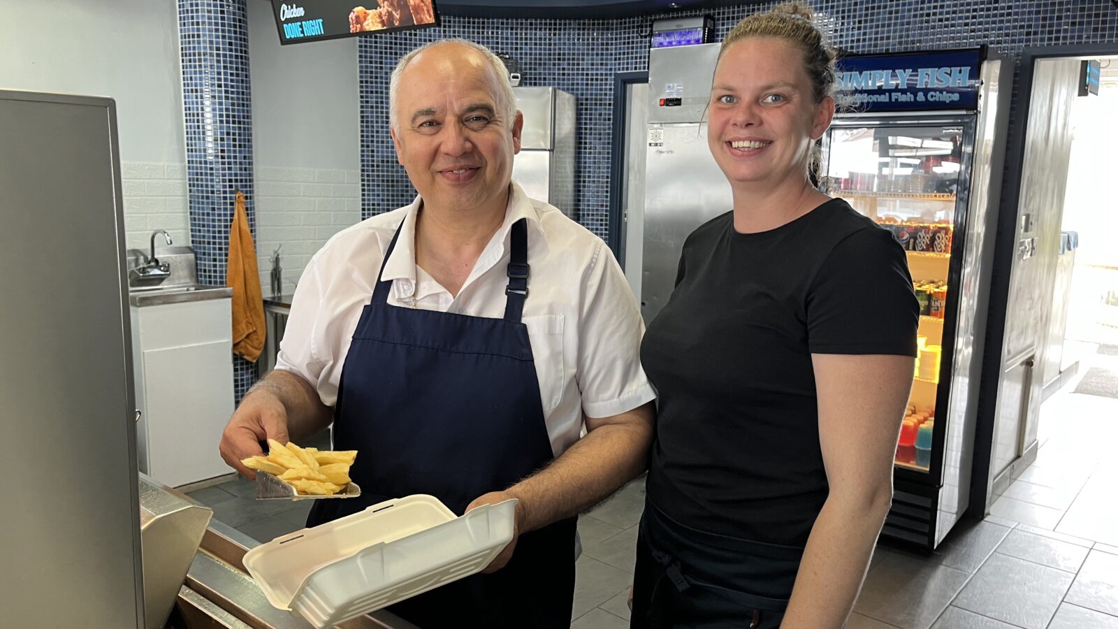 A man and a woman stand behind a fish and chip counter - the man is serving chips
