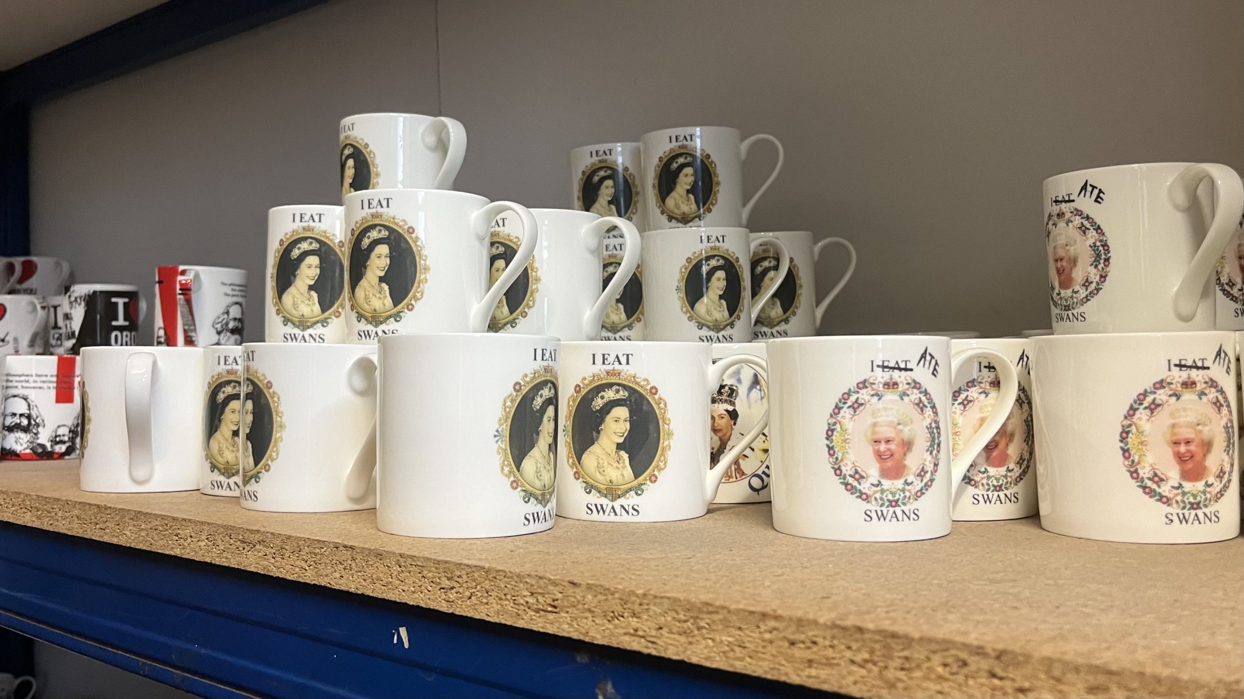 Mugs depicting an image of the late queen with the caption "i eat swans" are stacked on a shelf