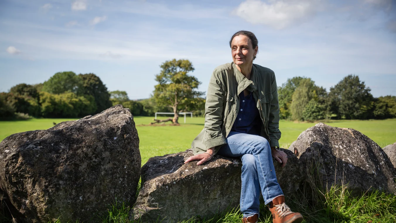 A woman sits on a rock in a field, with a copse of trees behind her 
