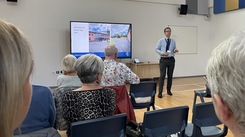 A teacher stands in front of a television in a large school hall.