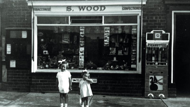 Black and white photo of two young girls standing outside a tobacconist and confectioners shop in the 1930s