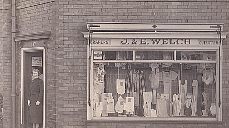 Black and white photo of a lady standing in the doorway of a small drapers shop