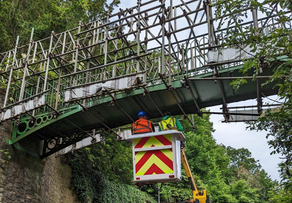 A small bridge, surrounded in scaffolding. Two men in a cherrypicker examine the underside