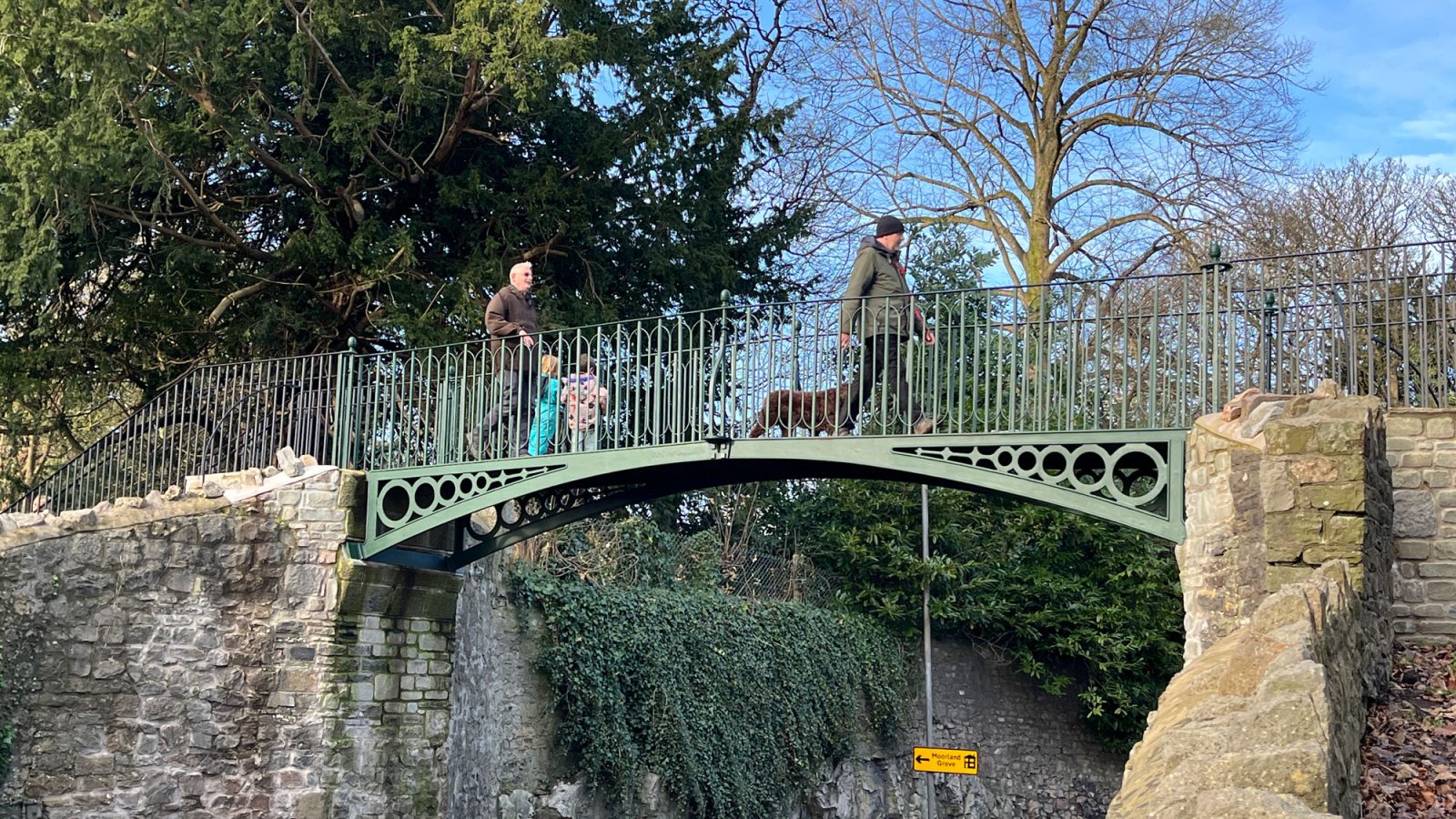 Pedestrians and dog walkers cross a small, green, iron bridge