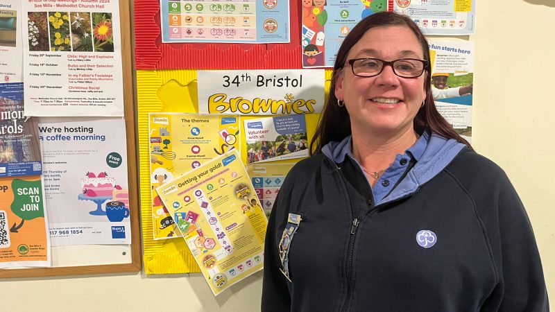 A women with long brown hair stands smiling in front of a notice board